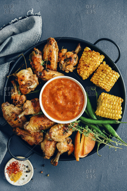 Grilled chicken wings are served on a black tray accompanied by grilled corn, green peppers, fresh thyme, peach slices and a bowl of sugar free bbq sauce in the middle photographed on a dark background from top view Chili flakes, turmeric and black peppercorns are on the side