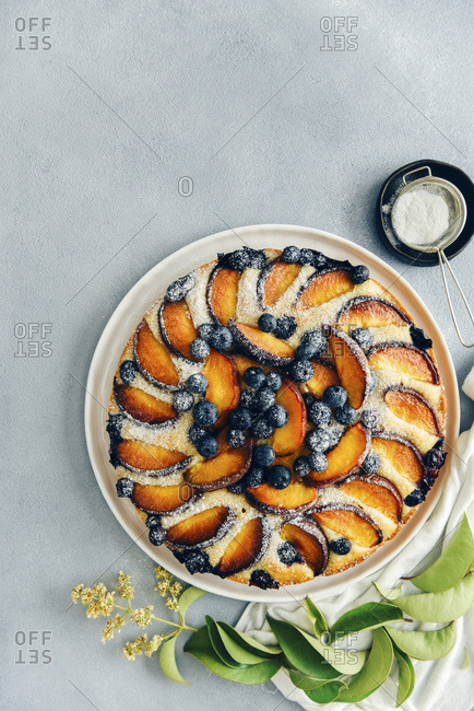Peach cake with blueberries dusted with powdered sugar photographed on a grey background from top view Green leaves and a mesh strainer with powdered sugar in a small black bowl accompany