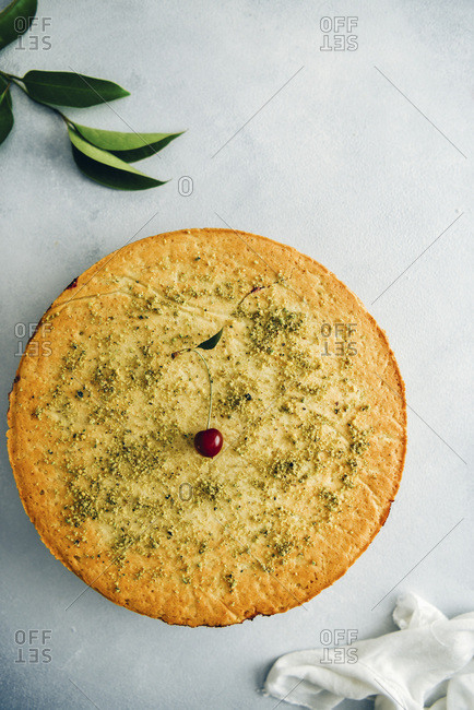 Cherry pie topped with a cherry and ground pistachio on grey background photographed from top view Green leaves and white linen accompany