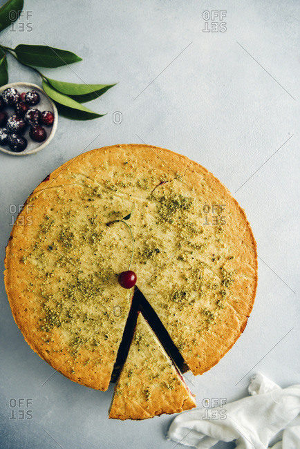 A cherry pie topped with a single cherry and ground pistachio with one slice photographed from top view Green leaves, pitted cherries on a small plate and white linen accompany
