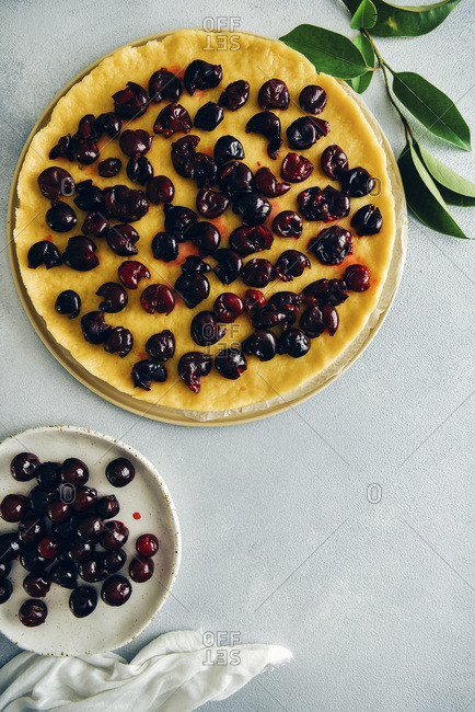 Pie dough topped with cherries, pitted cherries on a white plate and green leaves on grey background photographed from top view