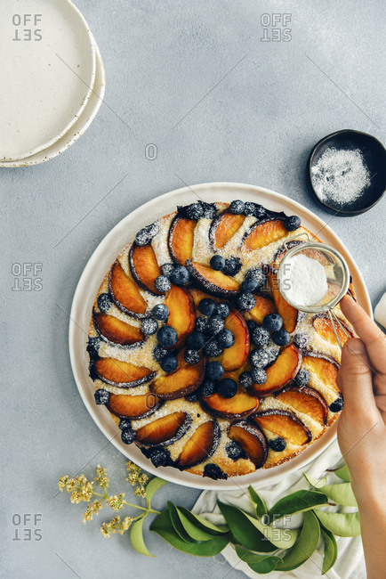 A woman sifting powdered sugar on a peach cake with blueberries photographed on a grey background from top view Green leaves, a black bowl with powdered sugar and mesh strainer and white ceramic plates accompany