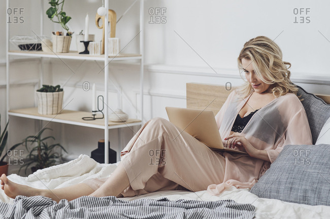 Beautiful sensual blonde Caucasian woman sitting on her bed in linen at morning and typing on laptop