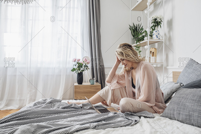 Beautiful blonde Caucasian woman sitting on her bed at morning and looking at laptop