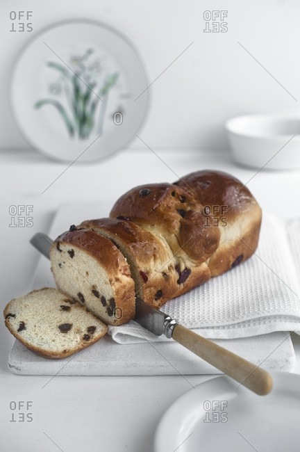 A fruit loaf, sliced, on a white chopping board with a knife and a cloth