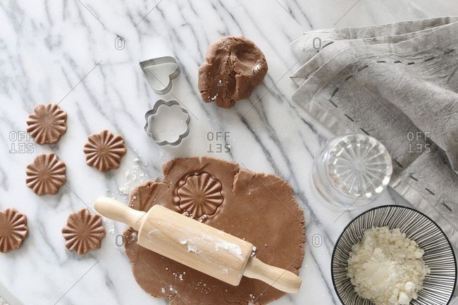 Gluten-free pastry rolled out with a rolling pin with cutters and a bowl of gluten-free flour on a marble surface