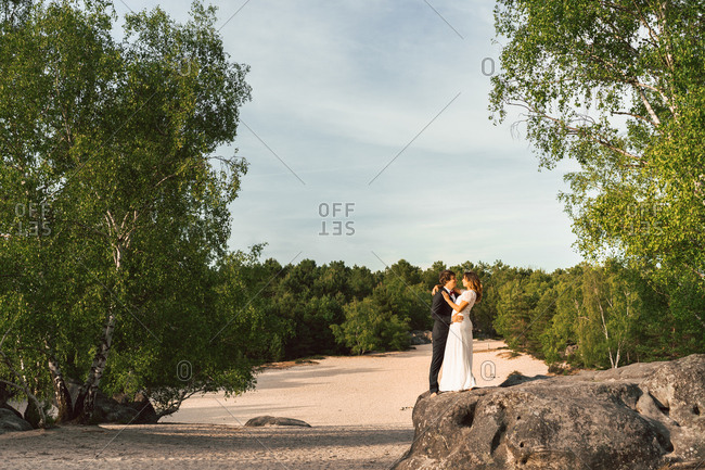 View at distance of couple in wedding gowns standing on rock and embracing happily against green trees and blue sky
