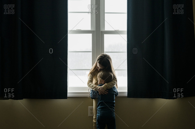 Young siblings in pajamas hug in front of large window