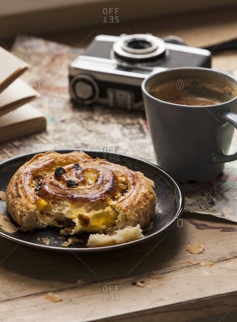 A cinnamon bun with raisins, a cup of coffee, a camera and a map on the wooden table