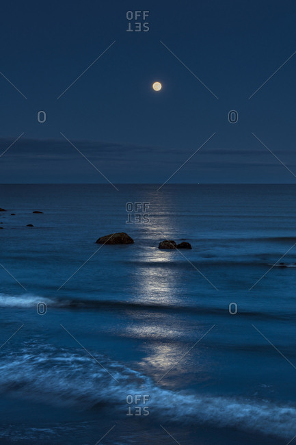 The rising full moon reflecting on the ocean at dusk with waves breaking into the shore.