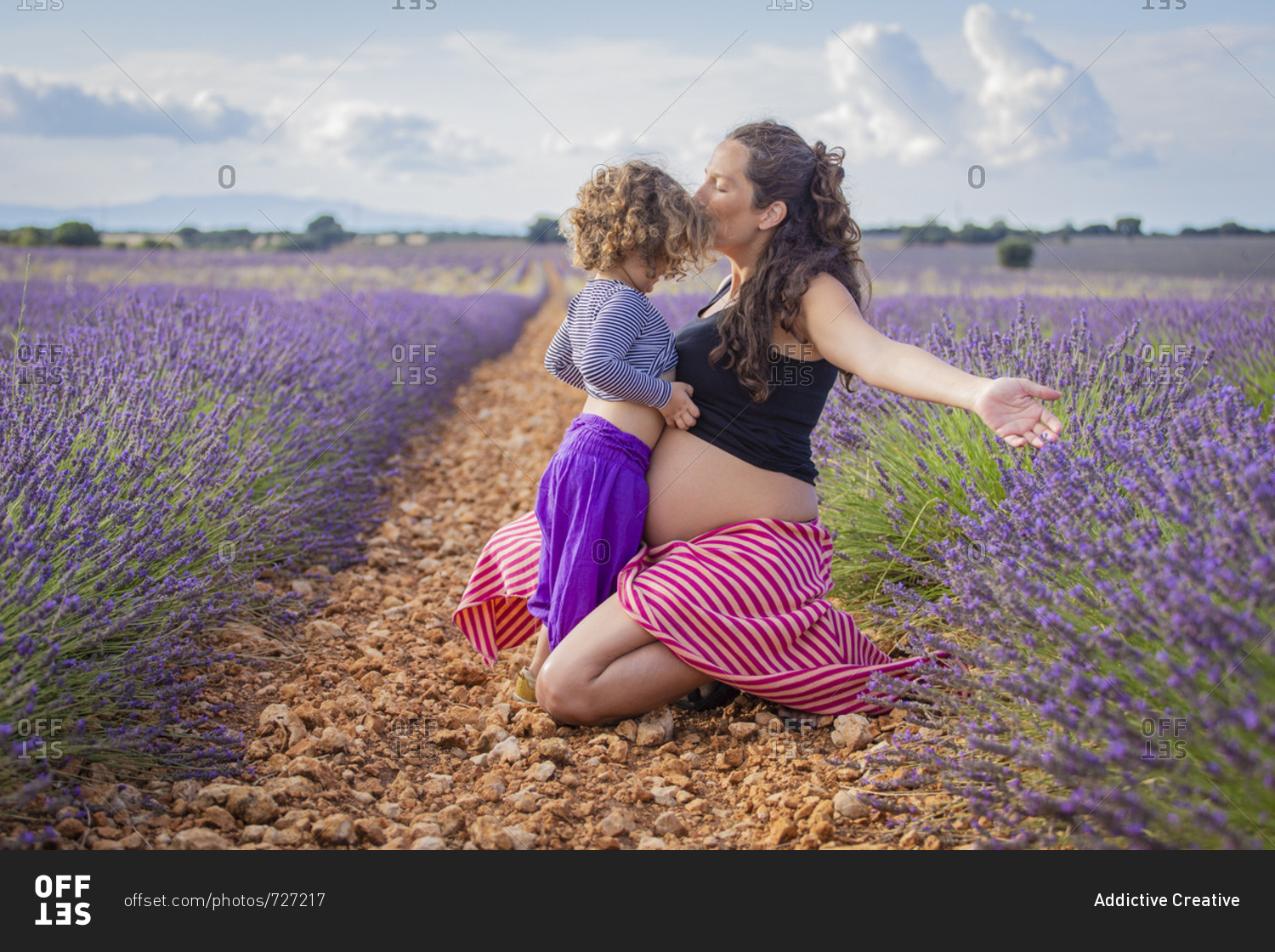 Side view of happy pregnant woman kissing little daughter while sitting on pathway in blooming lavender field