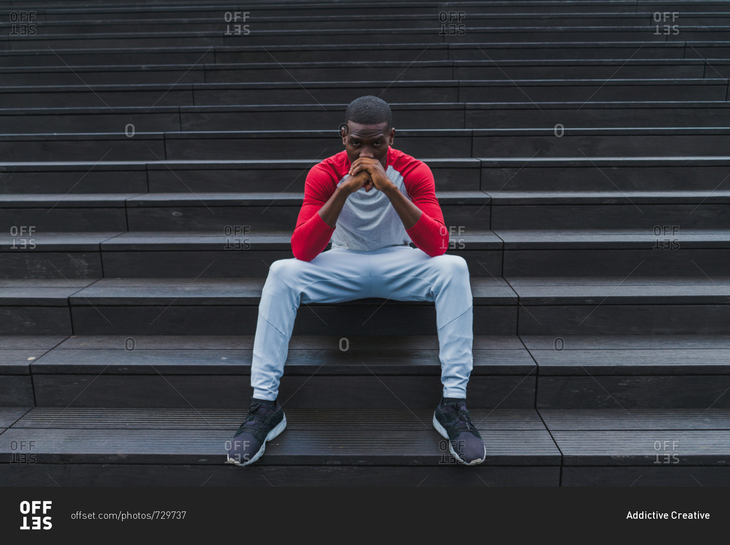 Handsome African-American resting man in sportswear sitting comfortably on grey wet stairs propping head with folded hands and looking at camera outdoors