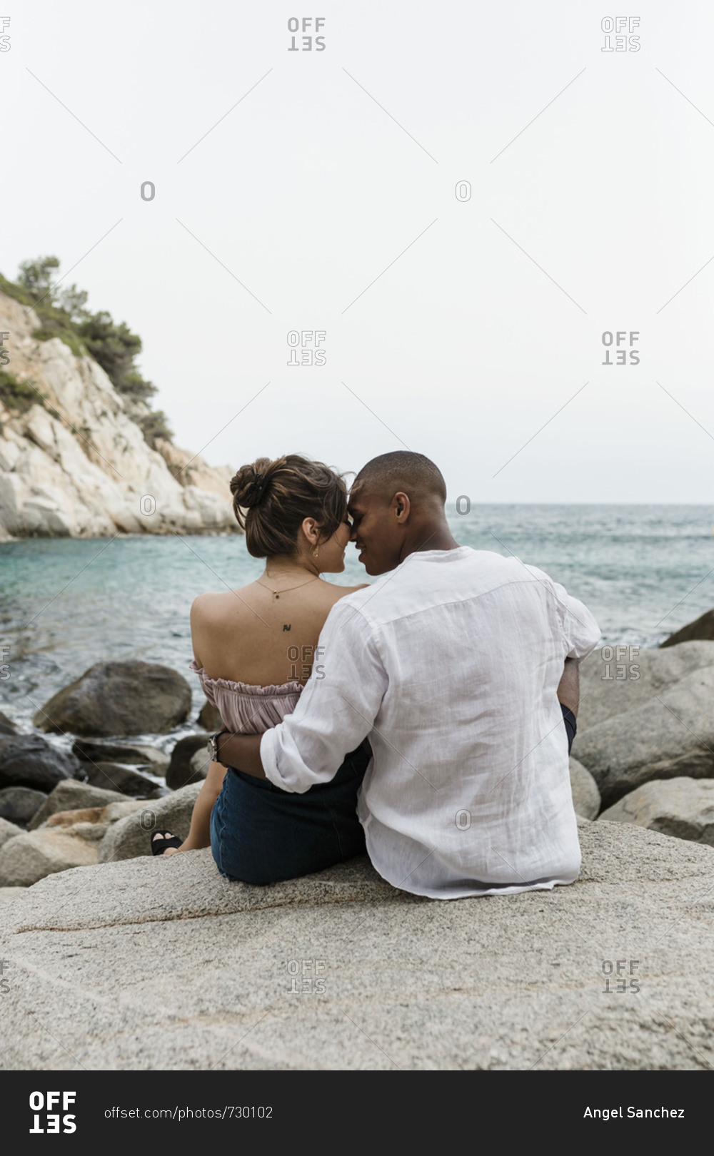 An interracial couple sits on the beach on a rock formation