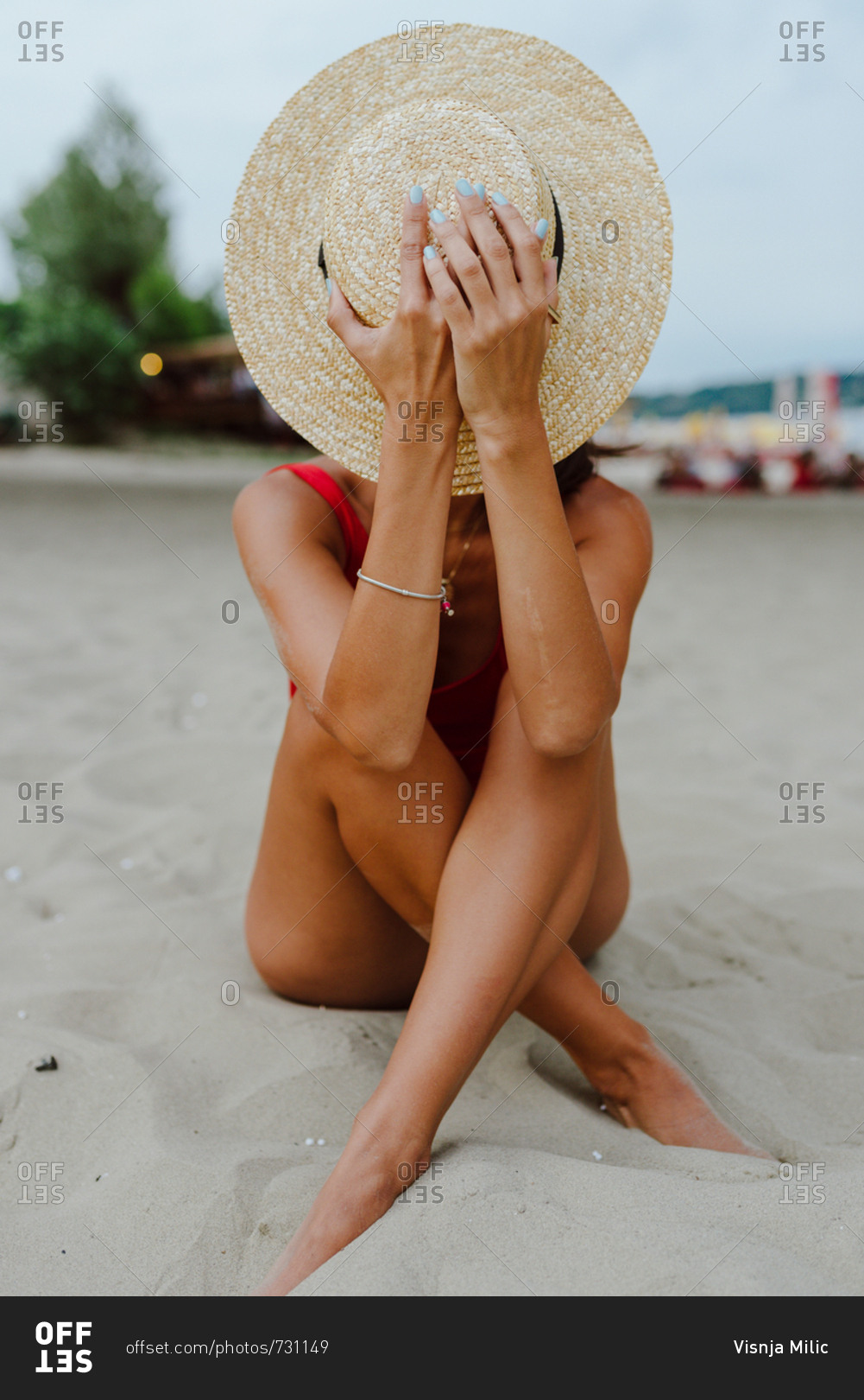 Woman in one piece red swimming suit sitting on the sand at the beach hiding her face with summer hat