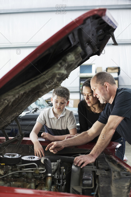 A senior caucasian male car mechanic talks to a caucasian mother and her son about a engine repair issue in a classic car repair shop.