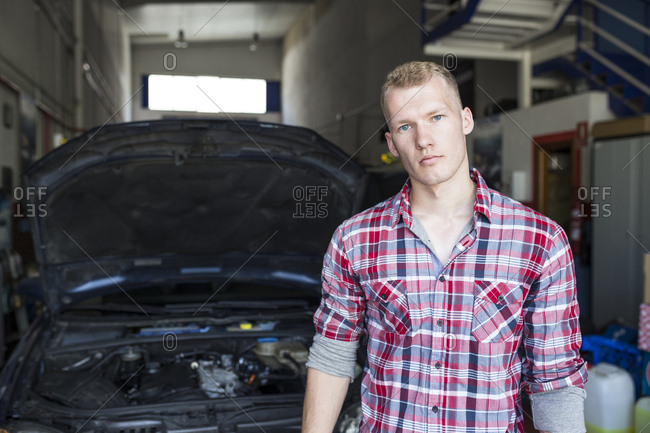 Man in plaid shirt standing near car with opened hood in repair shop
