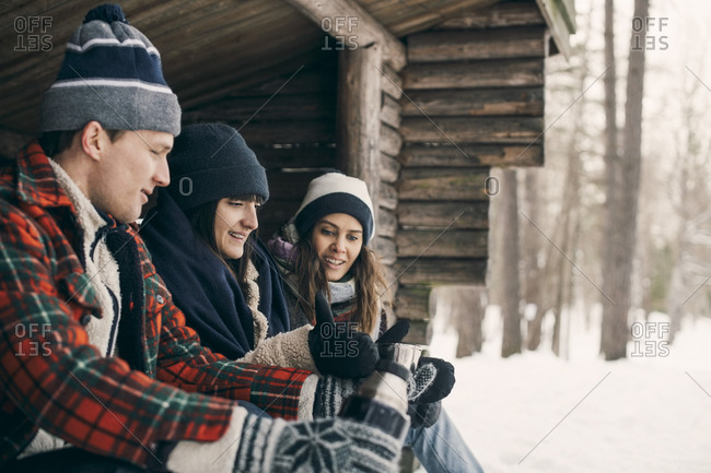 Man giving drink to female friends while sitting at log cabin during winter