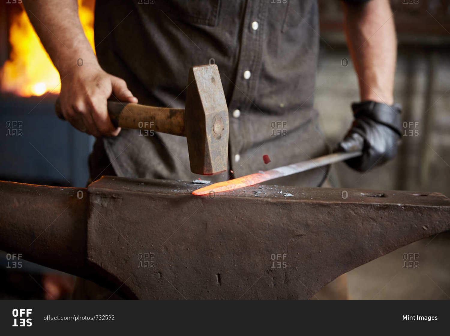 An artisan metal worker in ear protectors using a hammer to shape a red hot piece of metal on an anvil