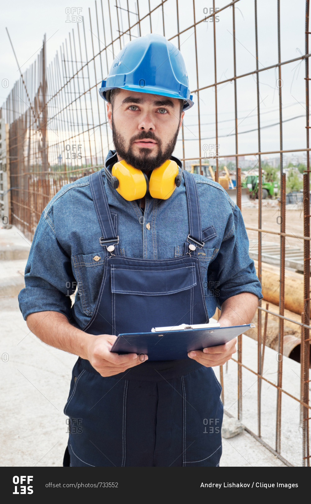 Portrait of confident bearded male general contractor in safety helmet standing at building site with clipboard and looking at camera