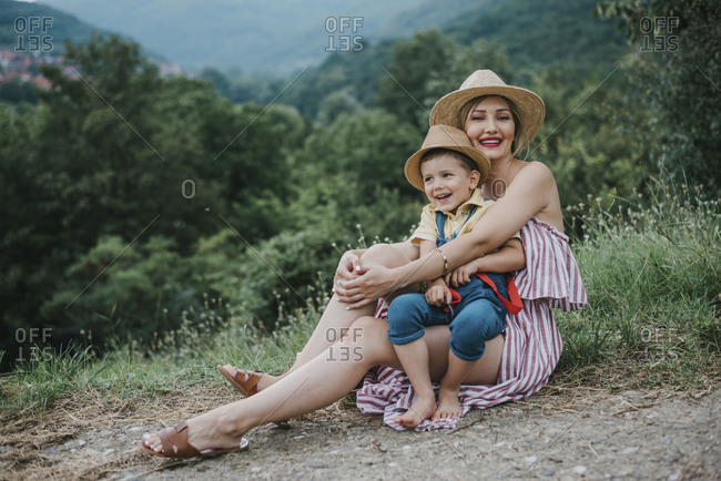 Young smiling mom sitting on a ground and holding her little kid among the grass in nature