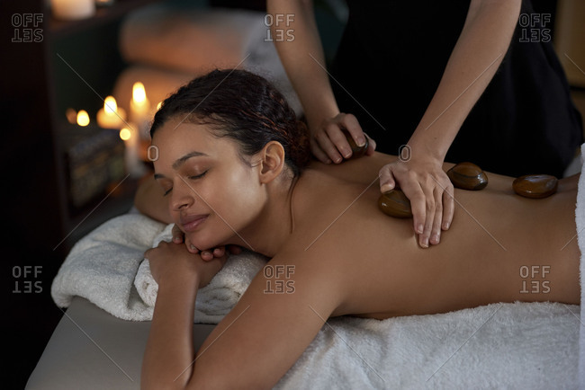 Beautiful woman enjoying relaxing hot stone massage by beauty therapist in  cozy treatment room lit with candles stock photo - OFFSET