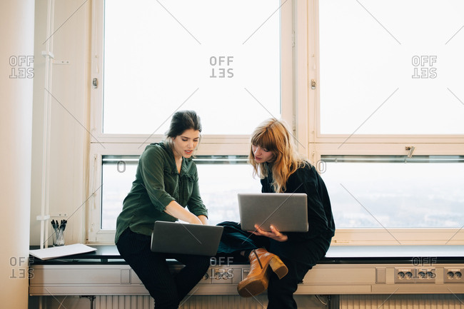 Confident female colleagues discussing over laptops while sitting on window sill at creative office