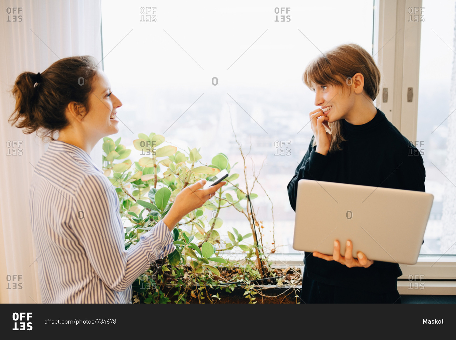 Smiling female colleagues standing with technologies by window at creative office