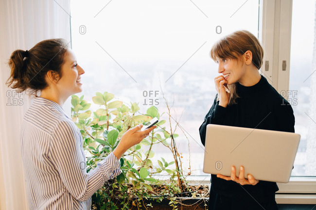 Smiling female colleagues standing with technologies by window at creative office