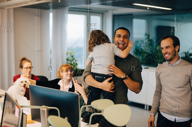 Cheerful businessman carrying son while standing amidst colleagues at creative office