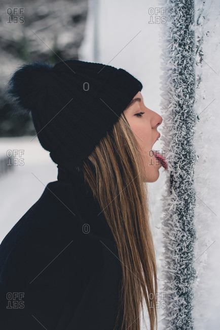 Blond lady in funny hat and black jacket eating snow from tree in winter forest side view