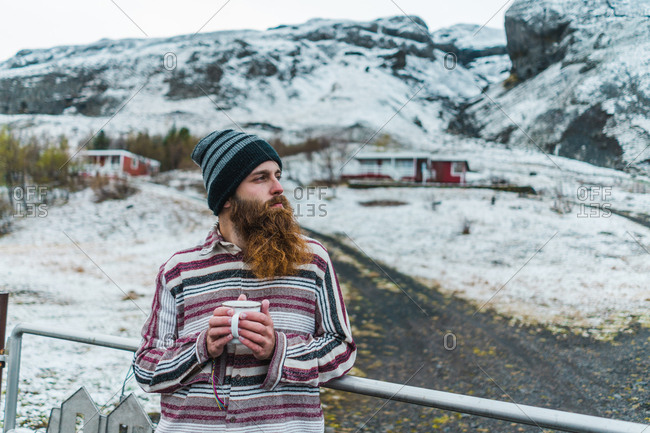 Attractive bearded man holding mug with hot drink and looking away while leaning on metal fence not far from small cottage in Icelandic countryside