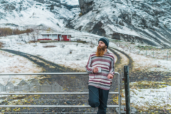 Attractive bearded man holding mug with hot drink and looking away while leaning on metal fence not far from small cottage in Icelandic countryside