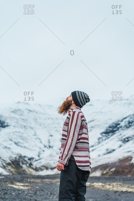 Attractive bearded man holding mug with hot drink and looking away while leaning on metal fence not far from small cottage in Icelandic countryside