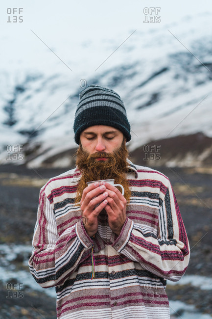 Attractive bearded man holding mug with hot drink and looking away while leaning on metal fence not far from small cottage in Icelandic countryside