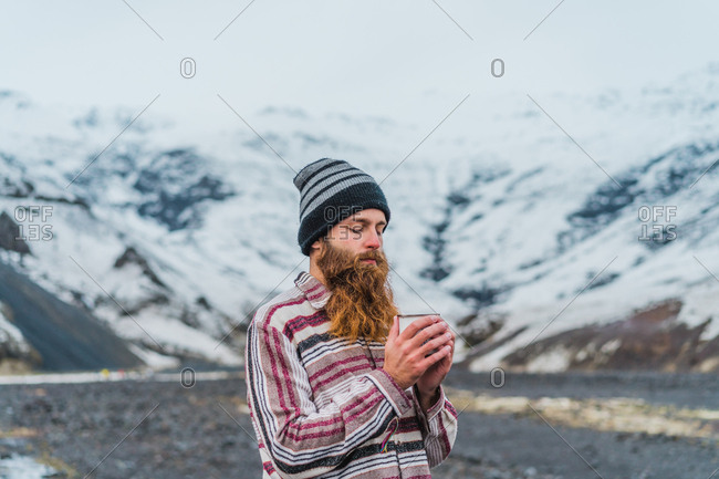 Attractive bearded man holding mug with hot drink and looking away while leaning on metal fence not far from small cottage in Icelandic countryside