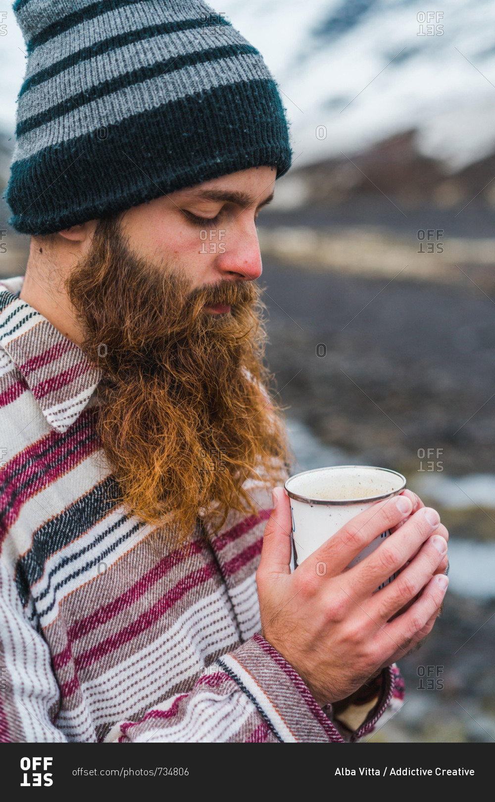 Attractive bearded man holding mug with hot drink and
looking away while leaning on metal fence not far from small
cottage in Icelandic countryside stock photo - OFFSET