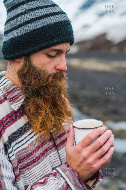 Attractive bearded man holding mug with hot drink and looking away while leaning on metal fence not far from small cottage in Icelandic countryside