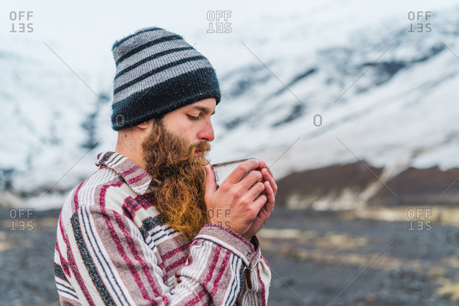 Attractive bearded man holding mug with hot drink and looking away while leaning on metal fence not far from small cottage in Icelandic countryside