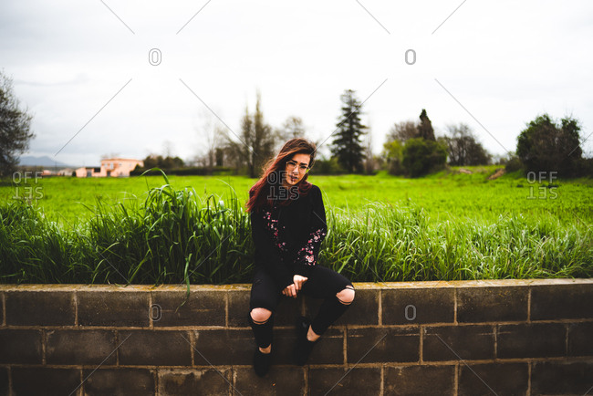 Side view of pretty woman in black sitting on brick wall with lush green  grass and gloomy sky on background. stock photo - OFFSET