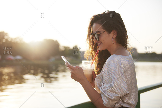 Smiling young woman with cell phone and takeaway drink at the riverside at sunset