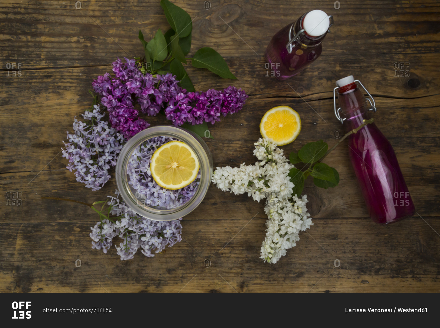 Two glass bottles of homemade lilac syrup and ingredients on wood