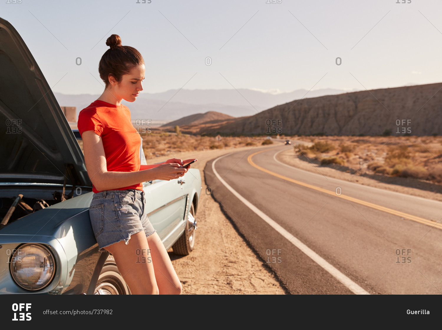 Young white woman leaning on broken down car using her phone