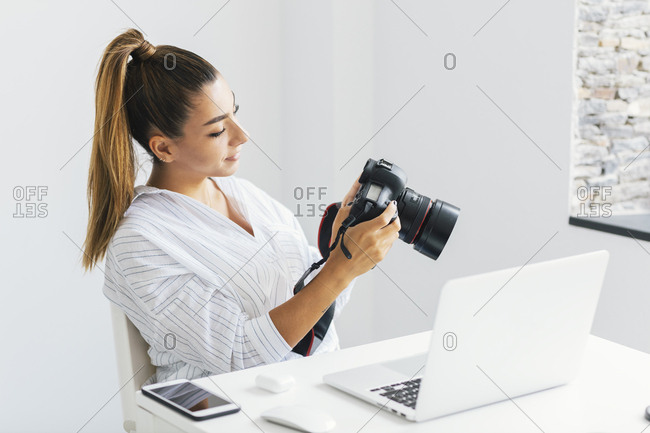 Woman holding and looking at digital camera while seated at desk in front of laptop computer