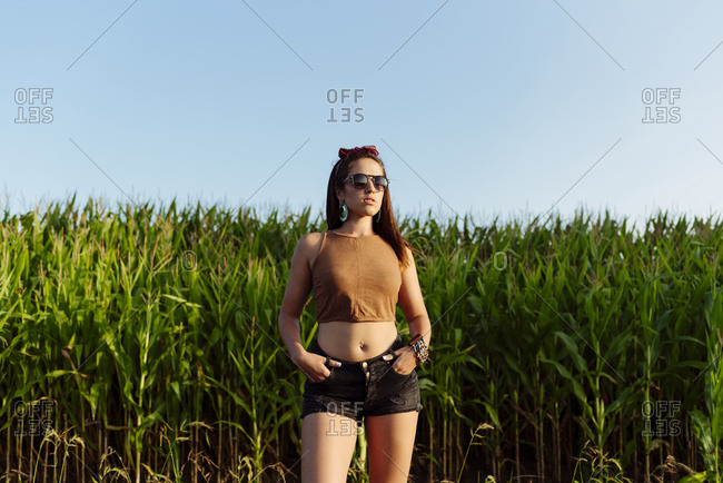 Beautiful brunette girl dressed in summer clothes with sunglasses standing near to a cornfield