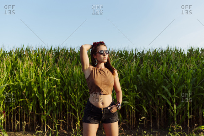 Beautiful brunette girl dressed in summer clothes with sunglasses standing near to a cornfield