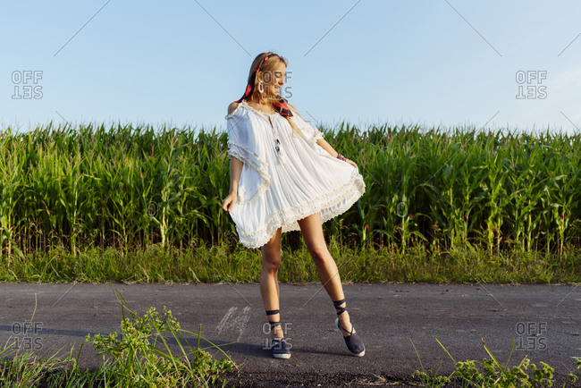 Pretty blonde girl with a white dress dancing on the road on a sunny day near to a cornfield