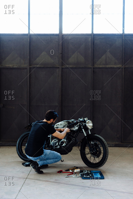 Young man kneeling to do maintenance on vintage motorcycle in empty warehouse
