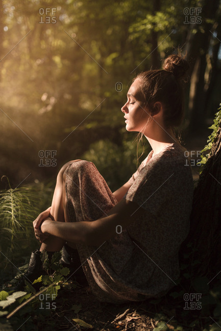 Young brunette with a hair bun sitting and relaxing under a tree sunlight through tree leaves