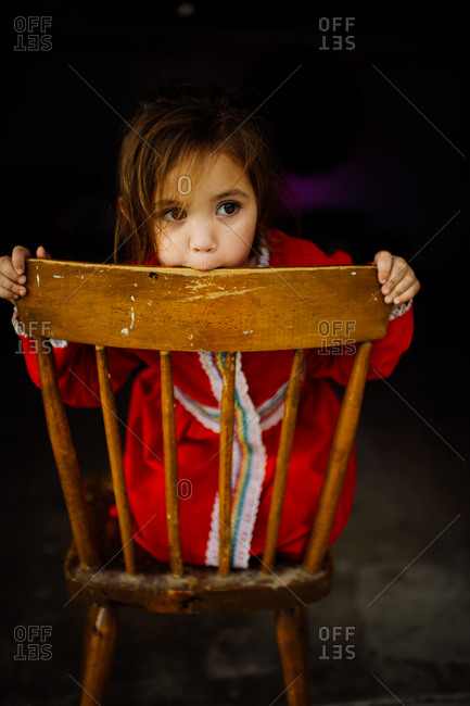 Toddler girl in red dress chewing on wooden chair