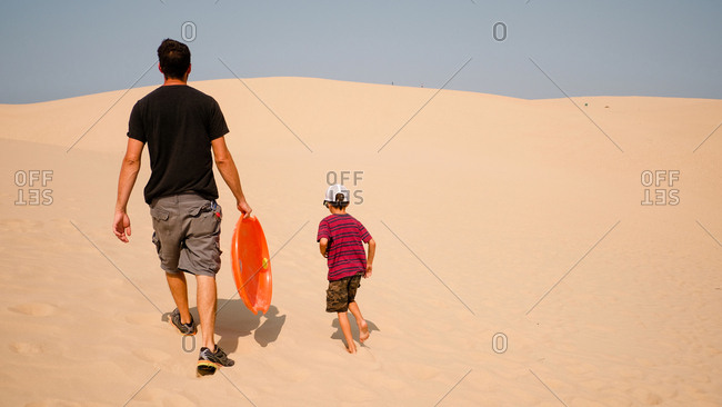 Father and son walking up sand dune with saucer for sledding